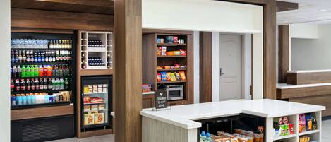 Una estación de alimentos y bebidas de autoservicio con almacenamiento refrigerado y en estanterías, un microondas y un cartel de bienvenida.