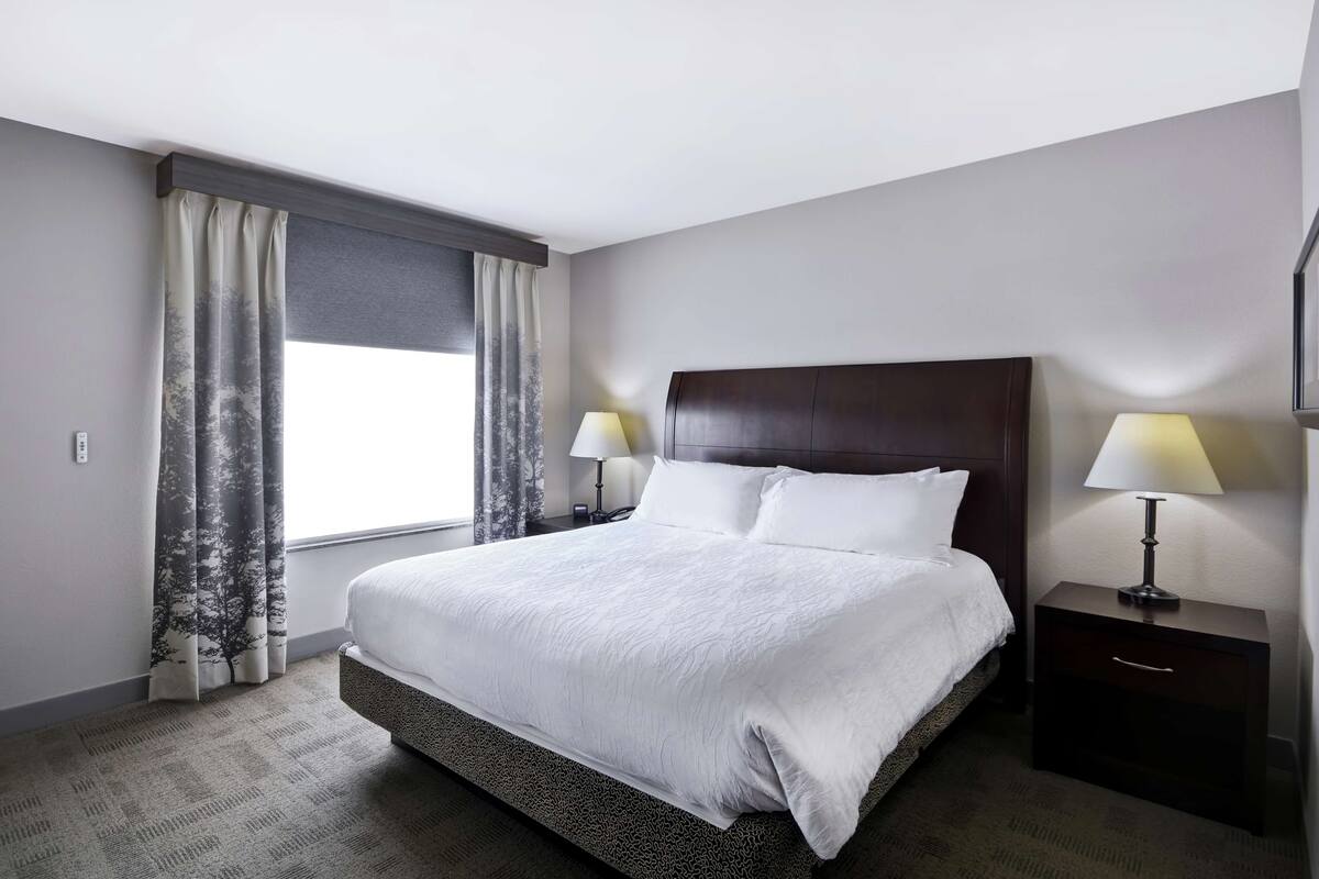 A hotel room with a large bed, two bedside lamps, a nightstand, and a window with curtains.