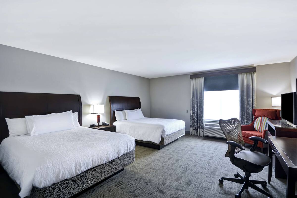 A hotel room with two beds, a desk, a chair, and a large window with curtains.