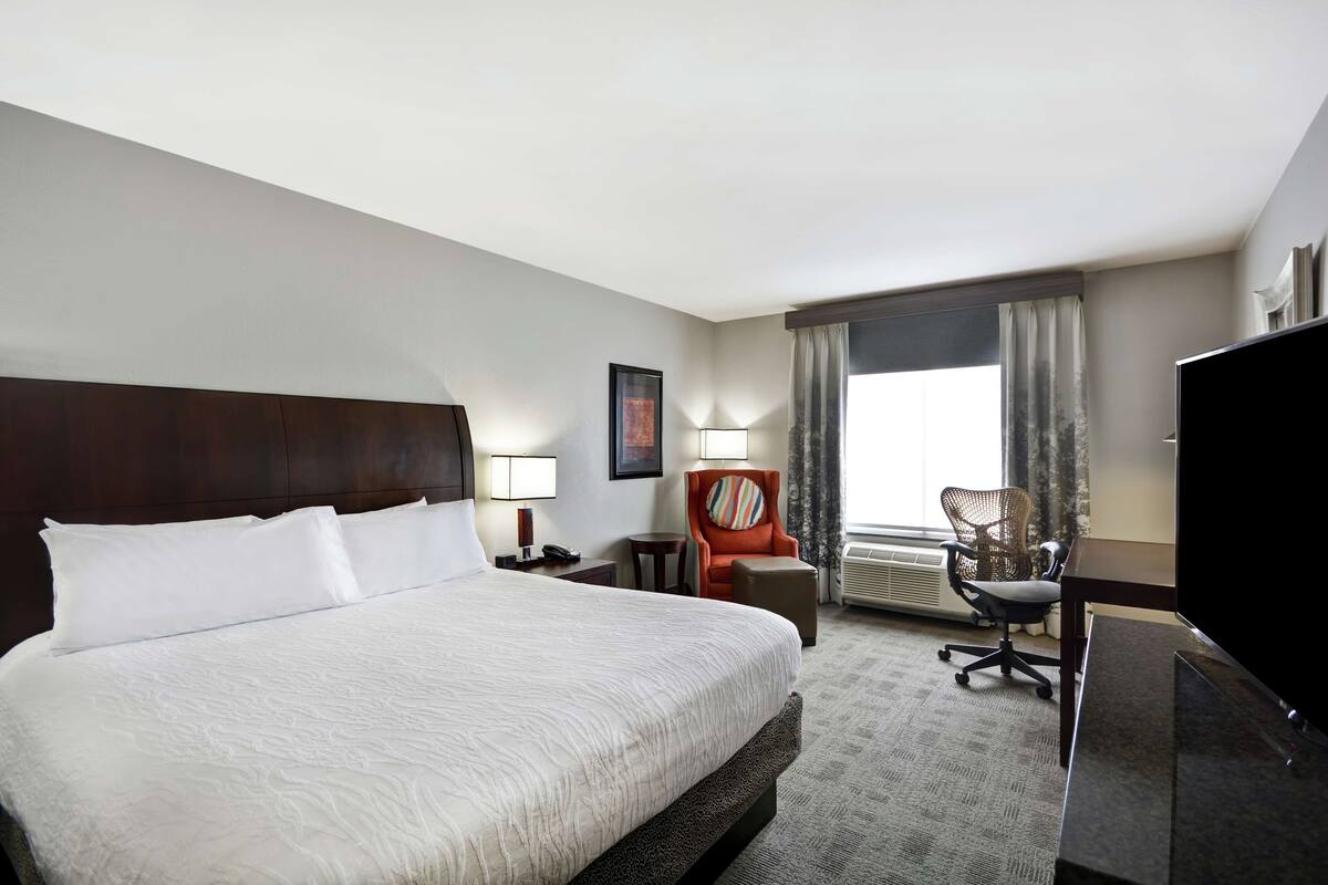 A hotel room with a large bed, a desk with a computer, a TV, a chair, and a window with curtains.
