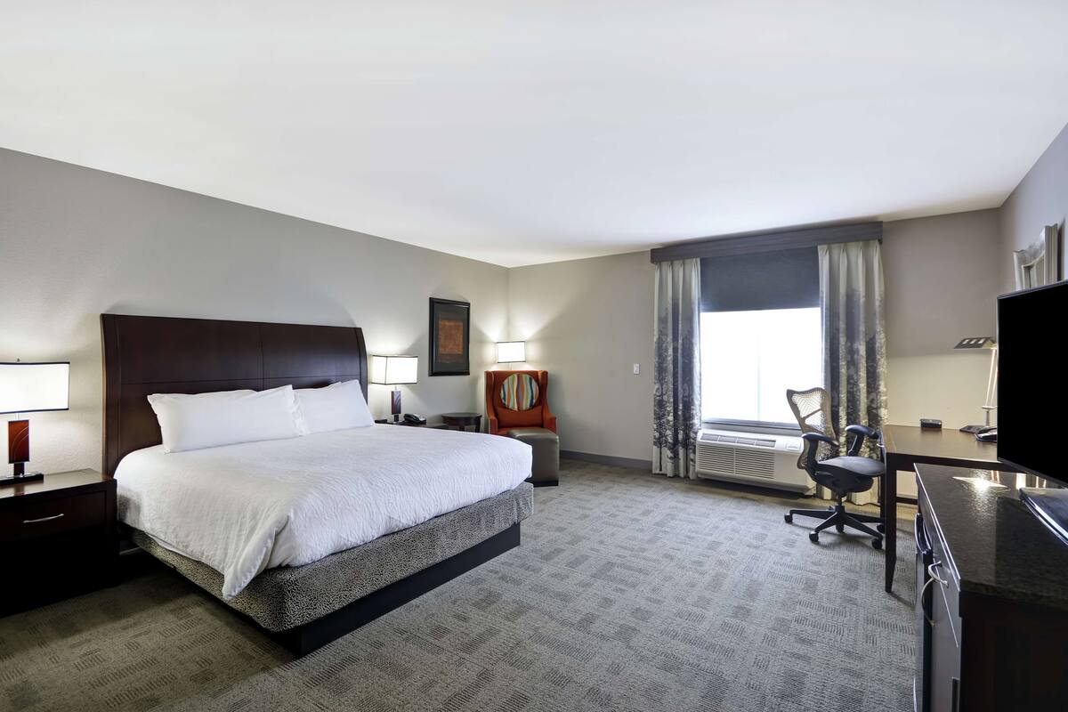 A hotel room with a large bed, a desk, a chair, a TV, and a window with curtains.