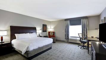 Habitación de hotel con una cama grande, un escritorio, una silla, un televisor y un ventanal con cortinas.