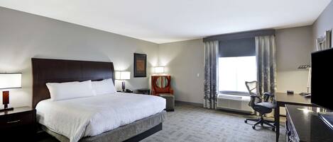 Ein Hotelzimmer mit einem großen Bett, einem Schreibtisch, einem Stuhl, einem Fernseher und einem Fenster mit Vorhängen.