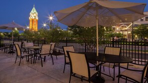 An outdoor patio with tables and chairs, a clock tower in the background, and string lights. - Hilton Garden Inn Toledo Perrysburg (Perrysburg)