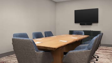 Ein Konferenzraum mit großem Tisch, sechs grauen Polsterstühlen, einem an der Wand montierten Flachbildfernseher und Teppichboden.