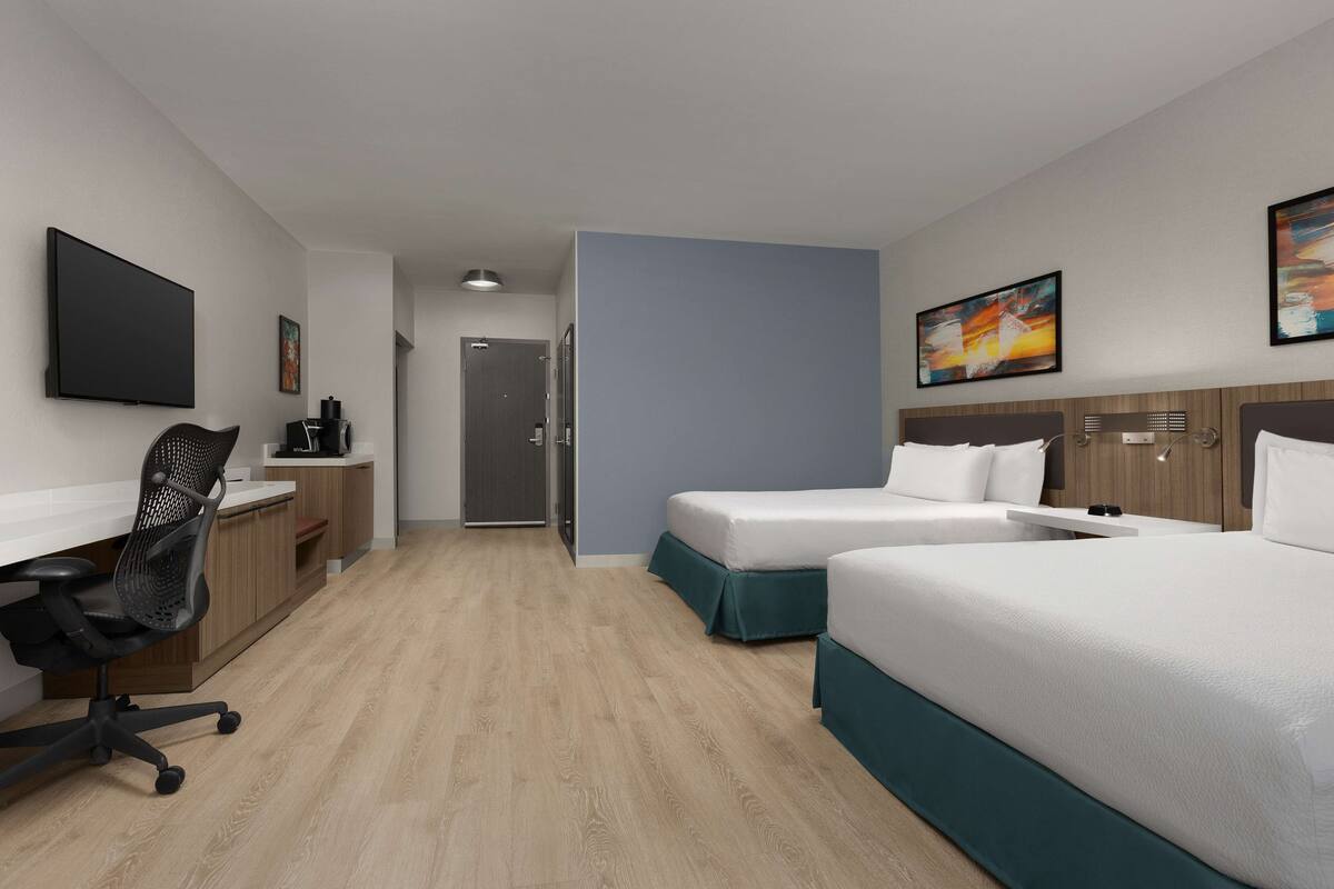 A hotel room with two beds, a desk, a chair, a TV, and artwork on the walls.