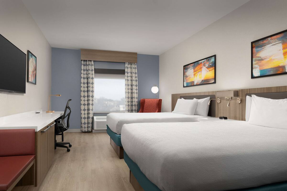A hotel room with two beds, a desk, a chair, and a view of the city.