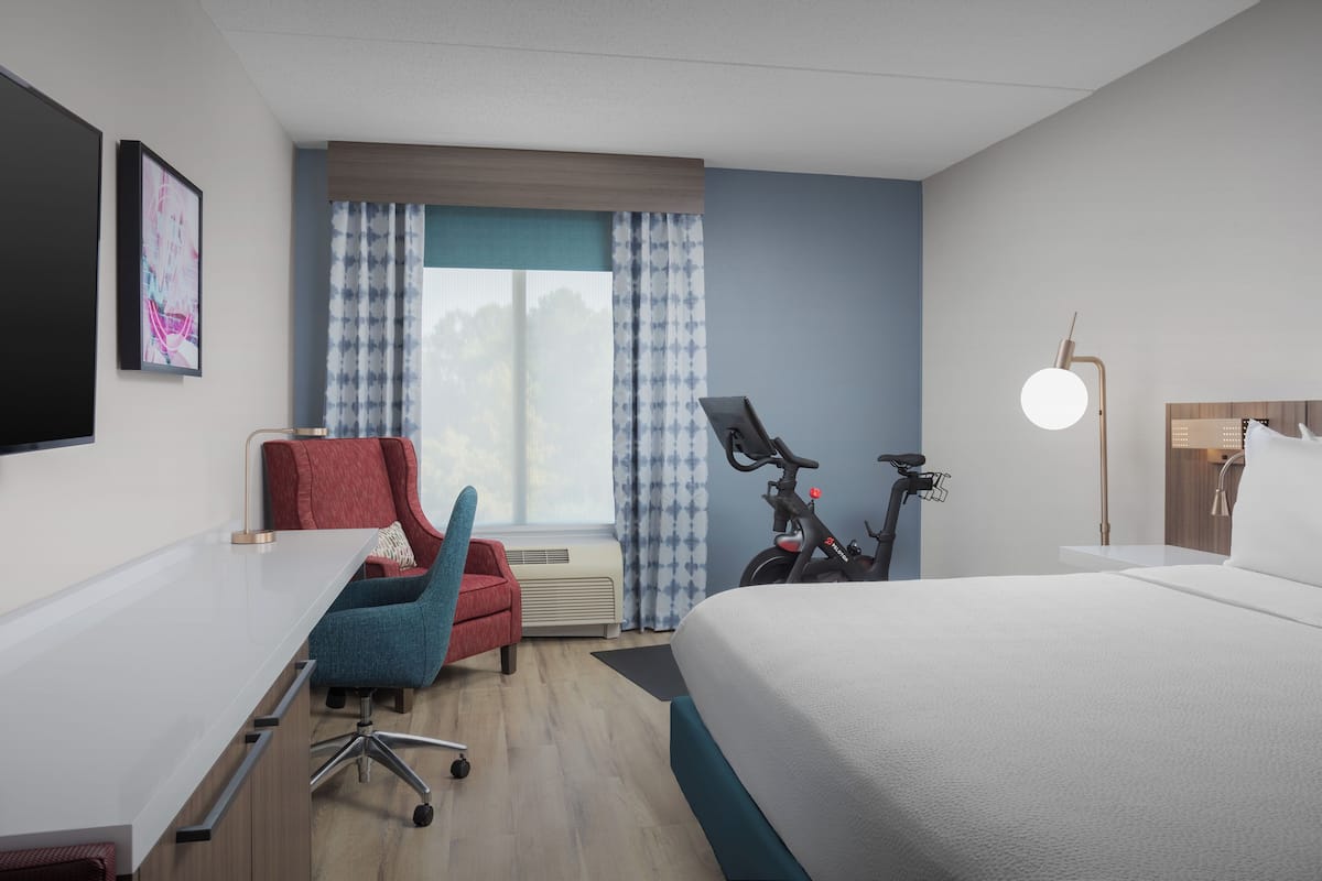 A modern hotel room with a large bed, a desk, a chair, a bicycle, and a window with curtains.