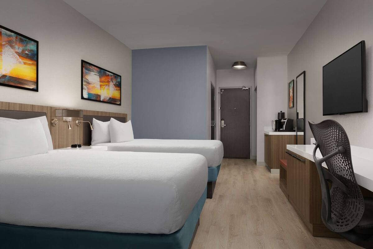 A hotel room with two beds, a desk, a chair, a TV, and artwork on the walls.