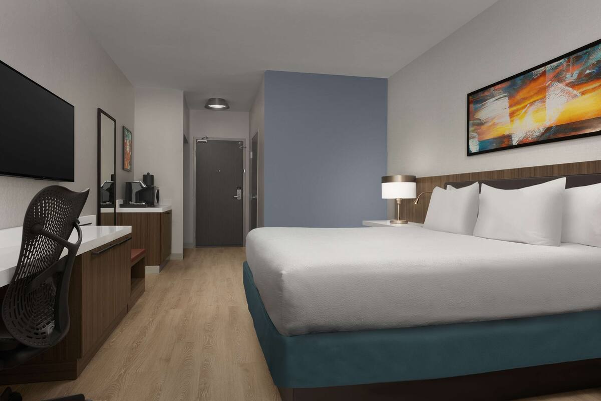 A hotel room with a bed, a desk, a chair, a TV, and a painting on the wall.