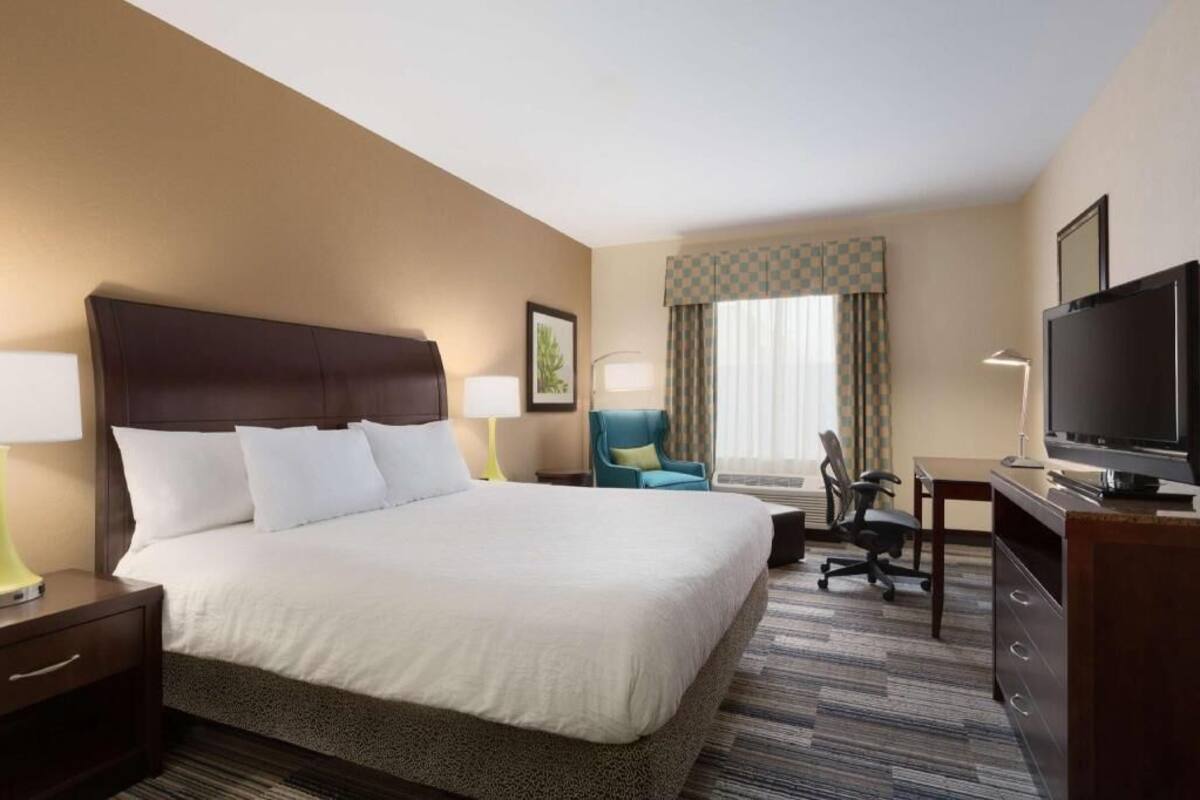 A hotel room with a large bed, a desk with a television, a chair, and a lamp.