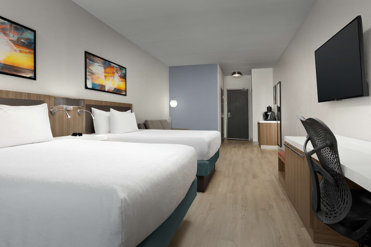 A hotel room with two beds, a desk, a chair, a TV, and artwork on the walls.