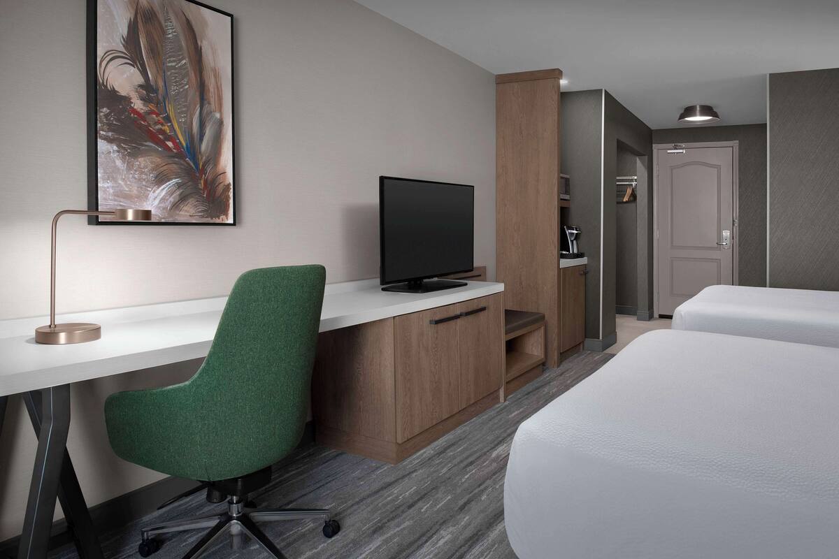Ein Hotelzimmer mit Schreibtisch, Fernseher, grünem Sessel, Bett und Tür.