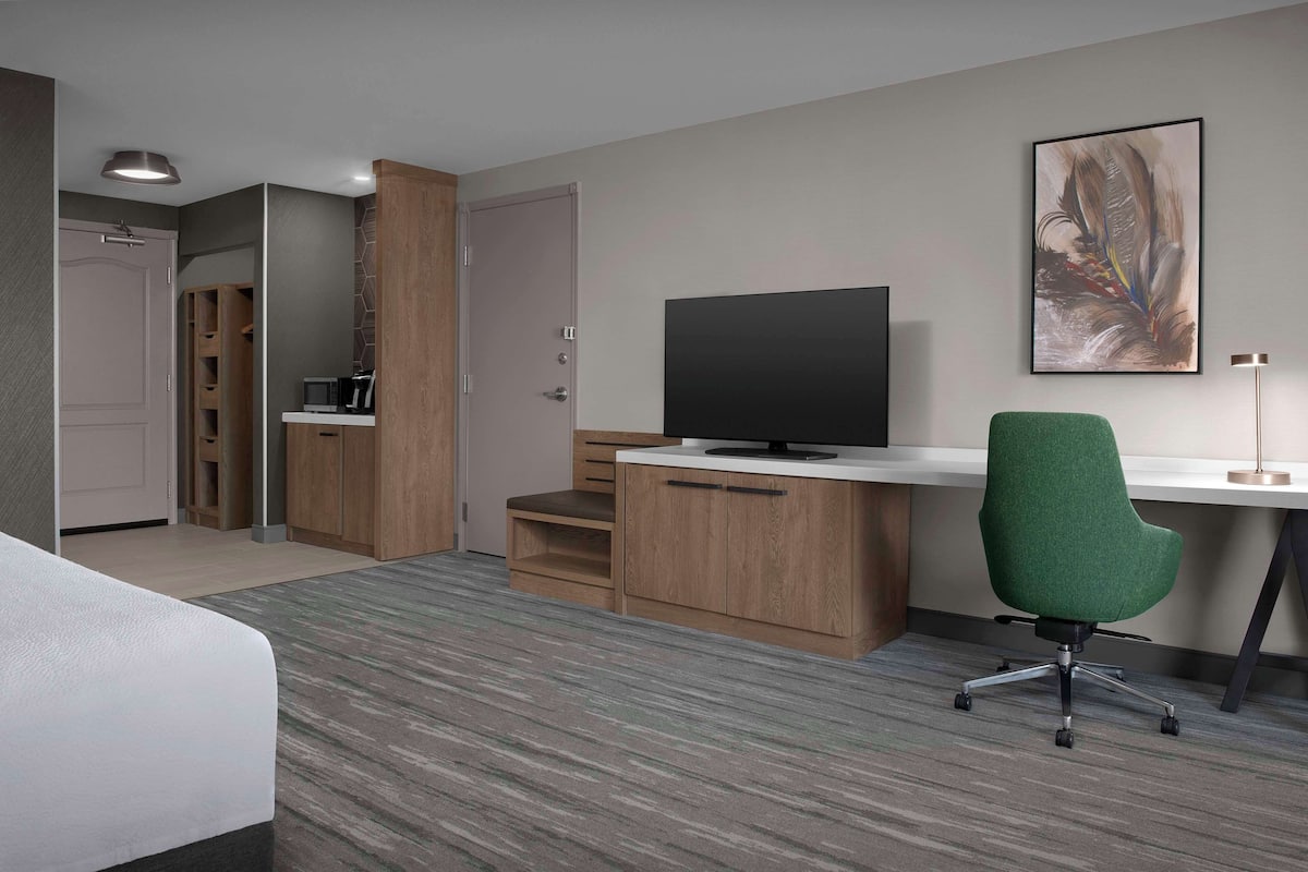 Ein Hotelzimmer mit einem Bett, einem Schreibtisch mit Fernseher, einem grünen Bürostuhl, einem Holzschrank und einem Gemälde an der Wand.