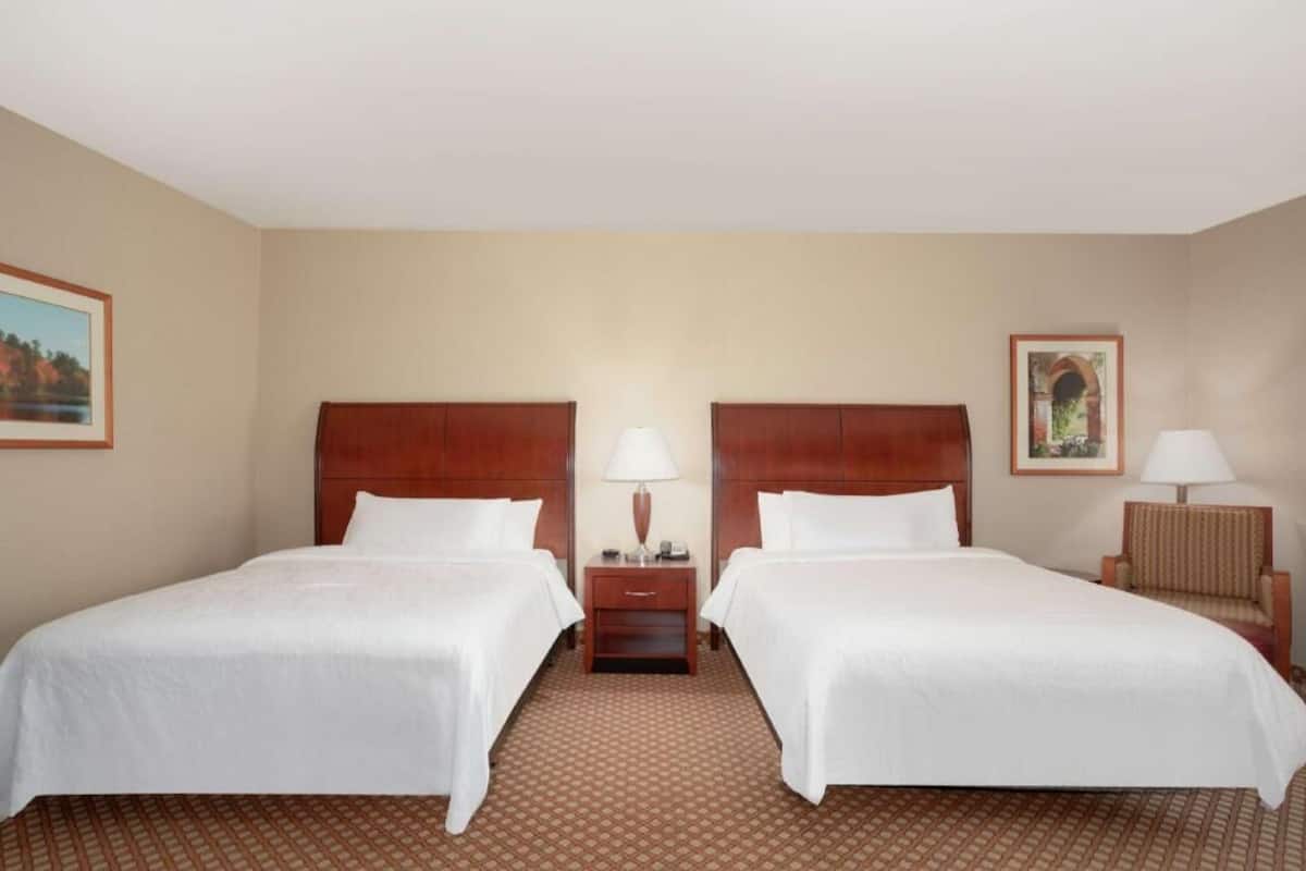 Ein Hotelzimmer mit zwei Betten, einem Nachttisch mit Lampe, einem Sessel und gerahmten Bildern an den Wänden.
