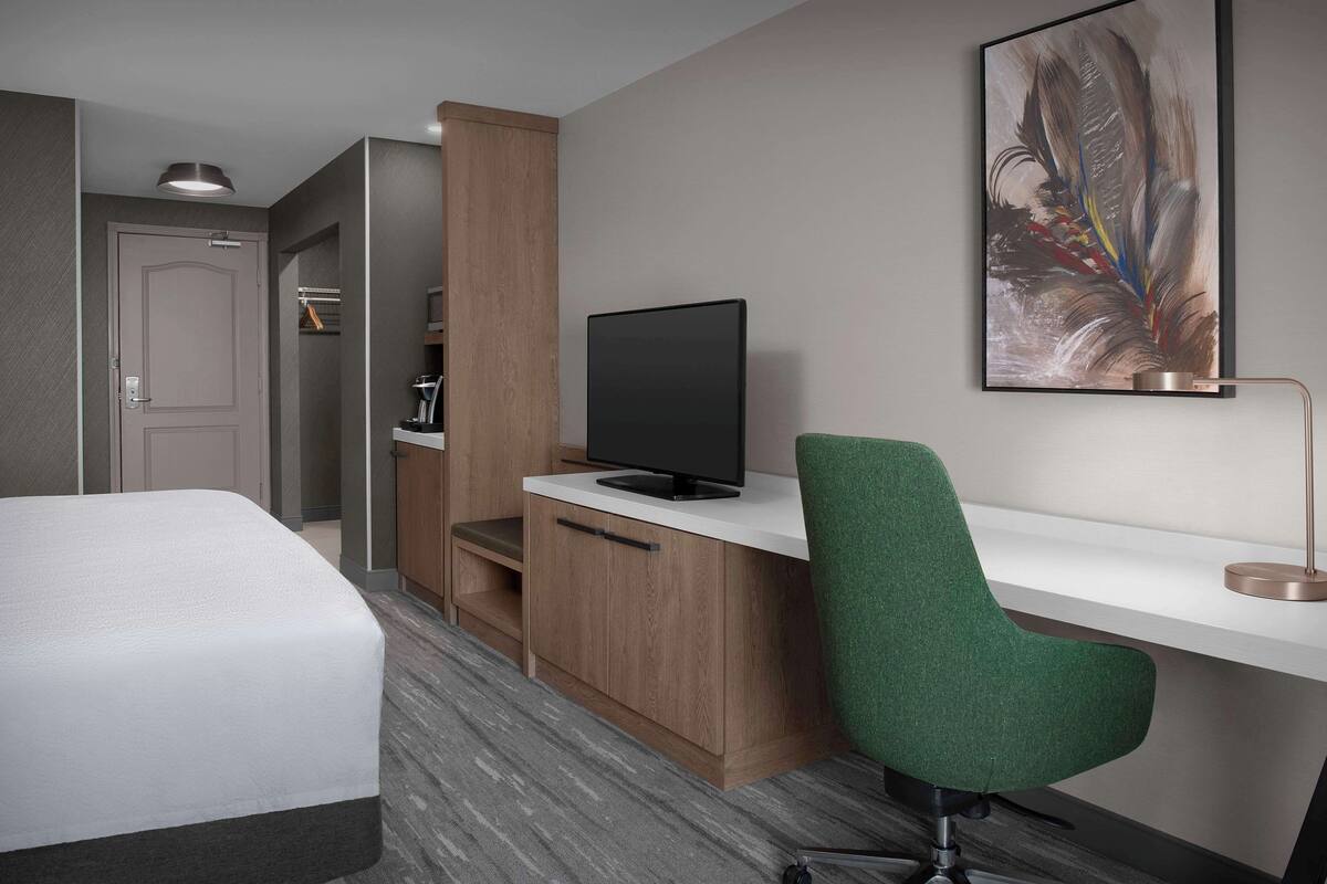 Ein Hotelzimmer mit Bett, Schreibtisch mit Fernseher, grünem Sessel, Kaffeemaschine und einem Bild an der Wand.