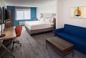 A hotel room with a bed, a desk with a chair, a blue sofa, a coffee table, and a TV.
