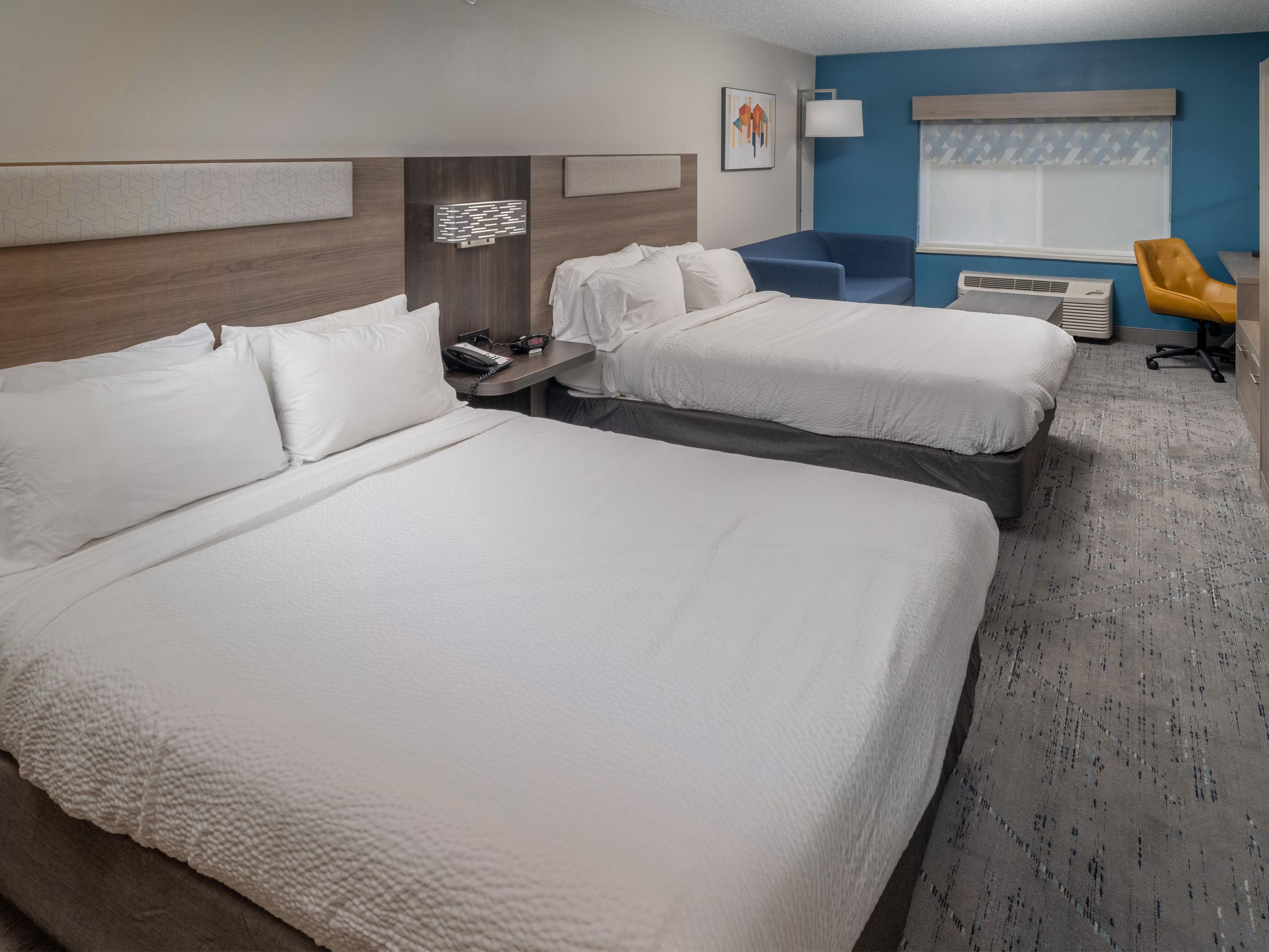 A hotel room with two beds, a desk, a chair, a lamp, and a window with blinds.