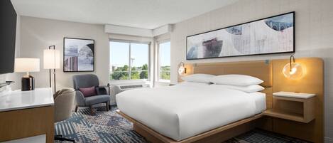 A hotel room with a large bed, a desk, a chair, a window with a view, and a wall-mounted artwork.