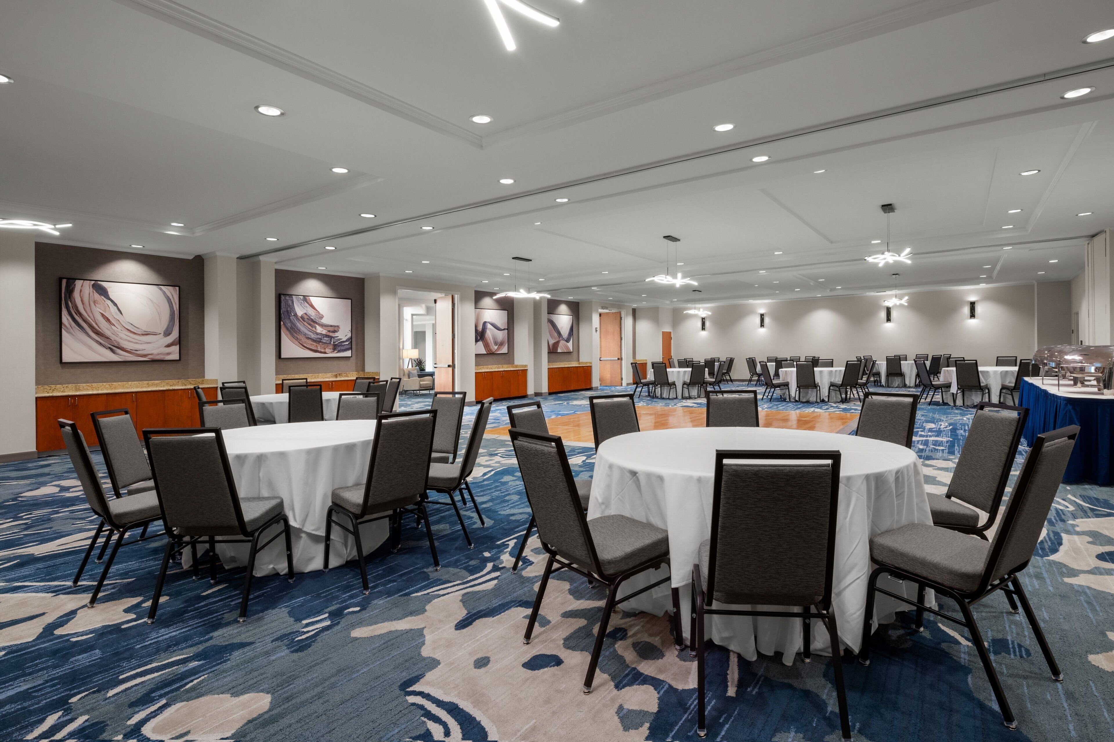 Une grande salle de conférence dotée de plusieurs tables rondes et de chaises, éclairée par des luminaires au plafond et agrémentée d’œuvres d’art sur les murs.