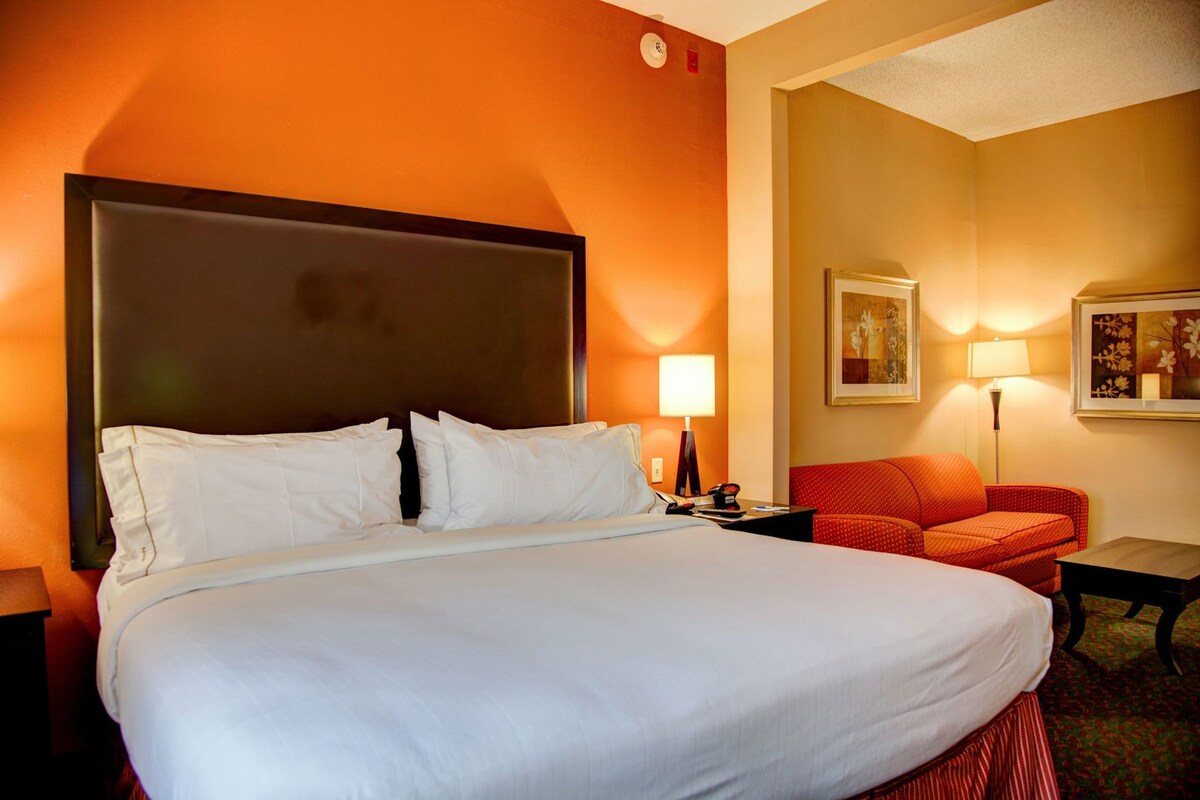 A hotel room with a large bed, a red armchair, a small table, and a lamp.