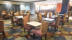 A hotel dining area with round tables and cushioned chairs, a patterned carpet, and a kitchen in the background. - Country Inn & Suites by Radisson, Tyler University Area (Tyler)