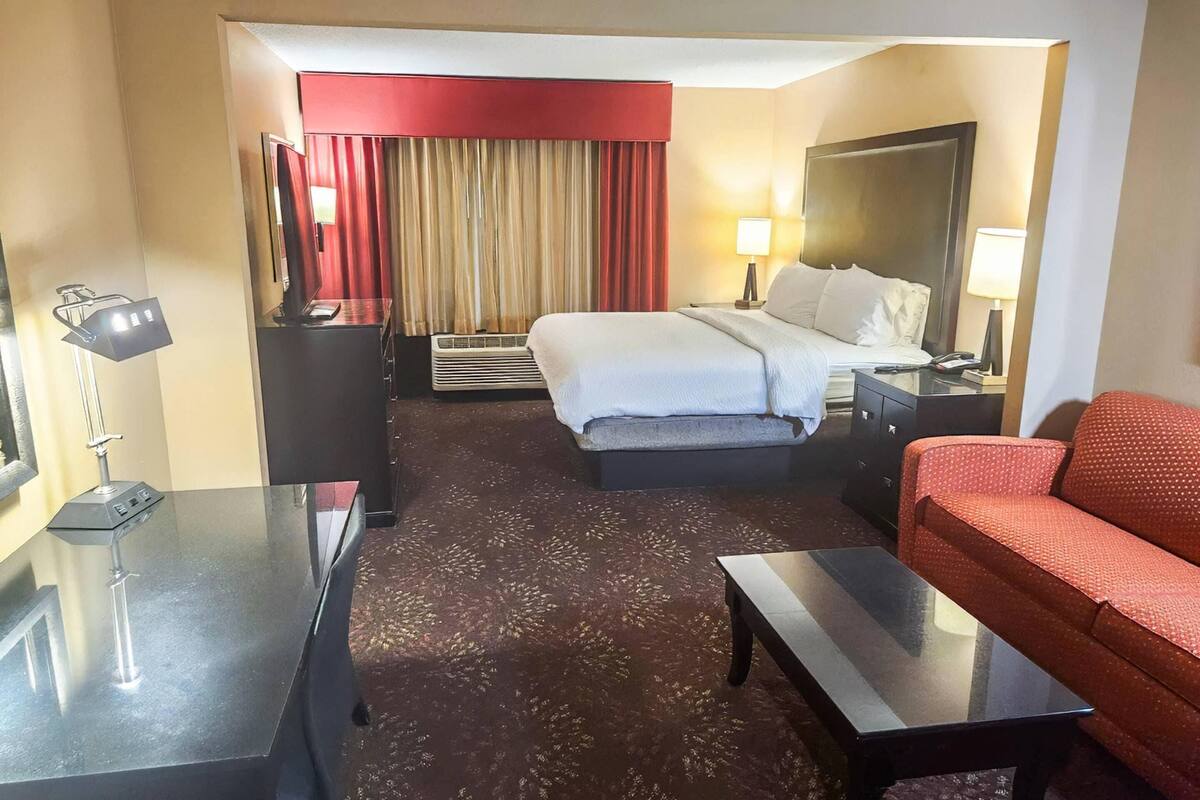 A hotel room with a bed, a sofa, a desk, a lamp, a mirror, and a red curtain.