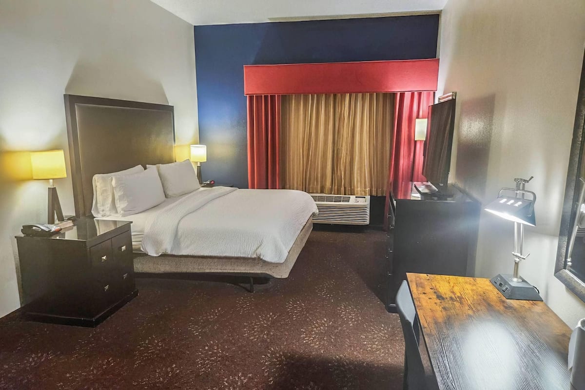 A hotel room with a bed, bedside tables, a desk, a television, and a window with curtains.