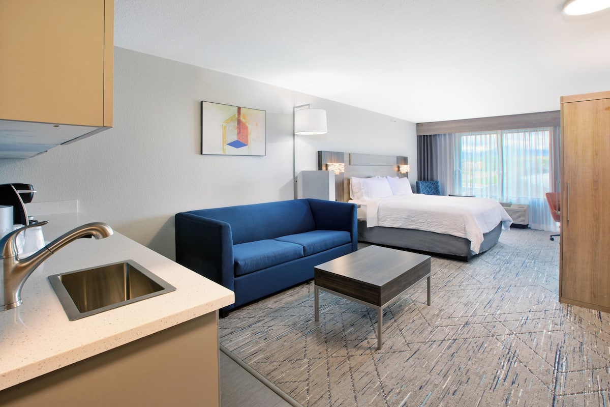 A hotel room with a kitchenette, a blue sofa, a bed with white linens, and a large window with curtains.