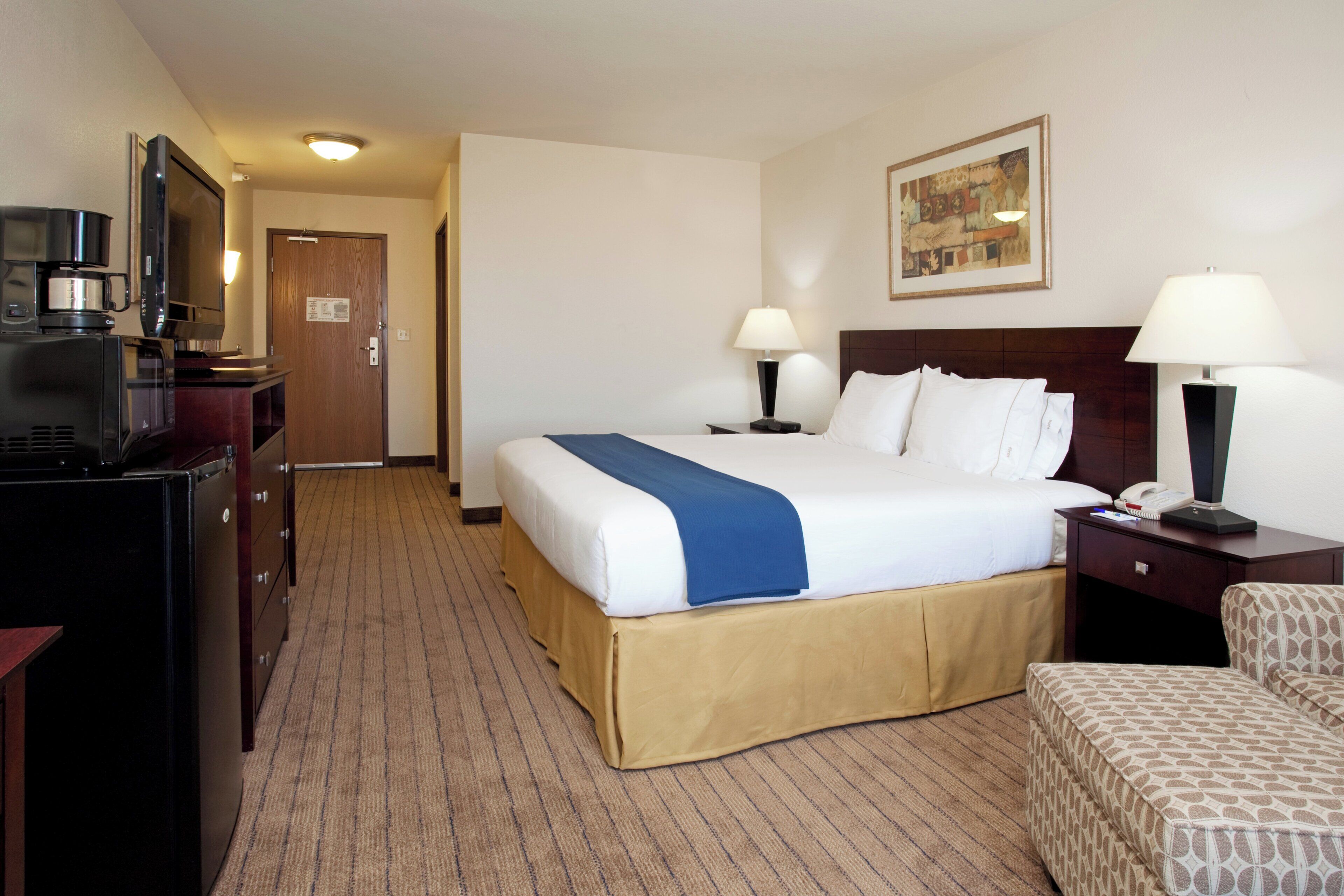 Habitación de hotel con cama, dos mesitas de noche con lámparas, escritorio con cafetera, una silla y puerta que da a otra habitación.