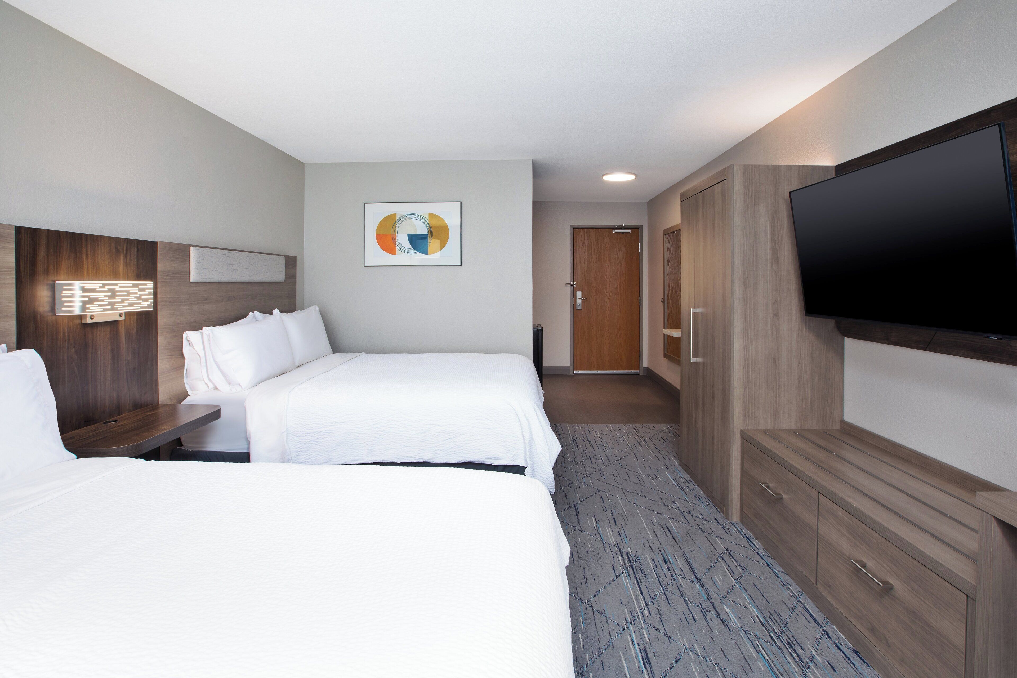 A hotel room with two beds, a flat-screen TV, a wooden wardrobe, and a framed abstract art piece on the wall.