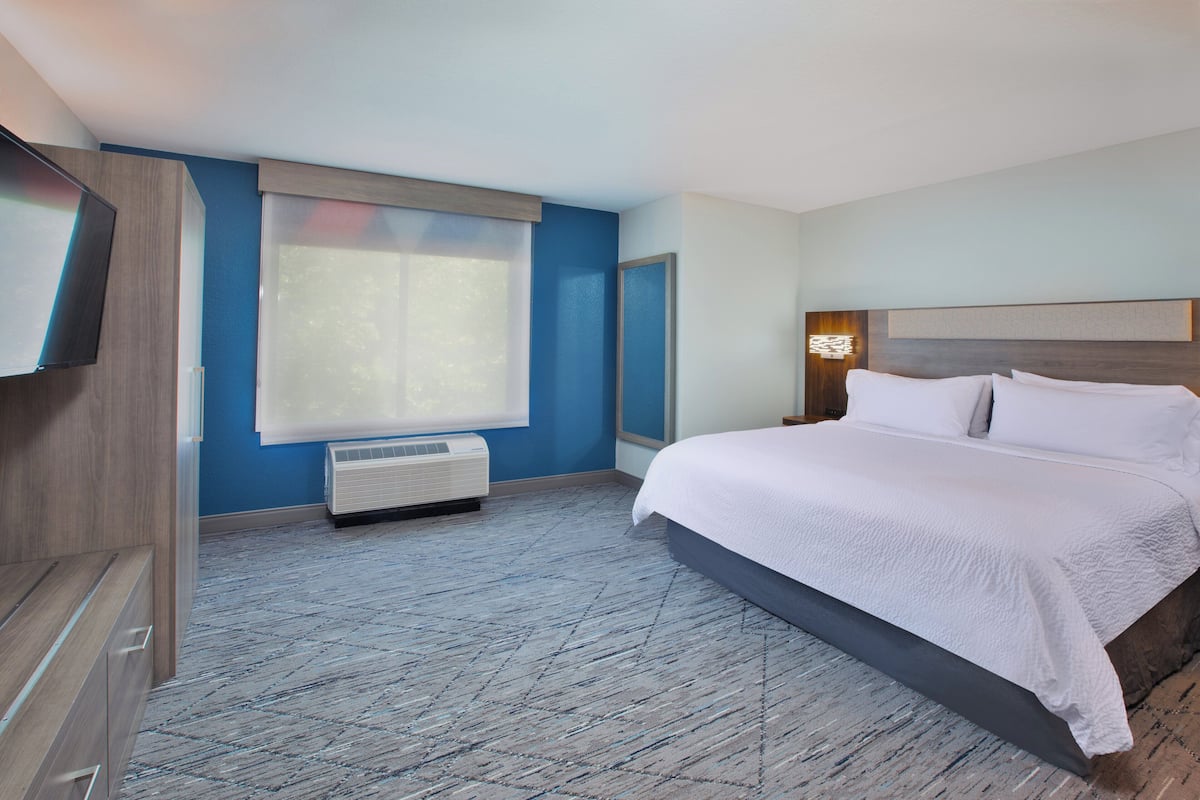 A hotel room with a large bed, a TV mounted on the wall, and a window with blinds.