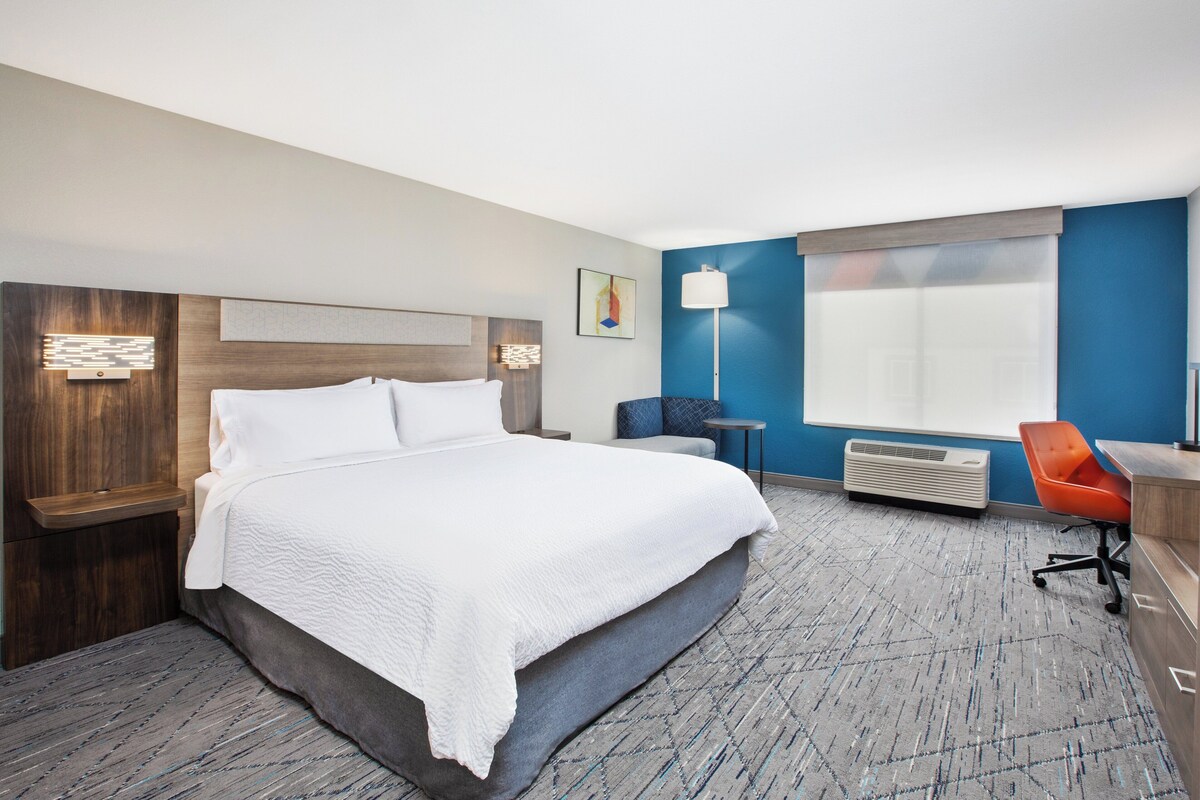 A hotel room with a large bed, a desk, a chair, a standing lamp, and a window.