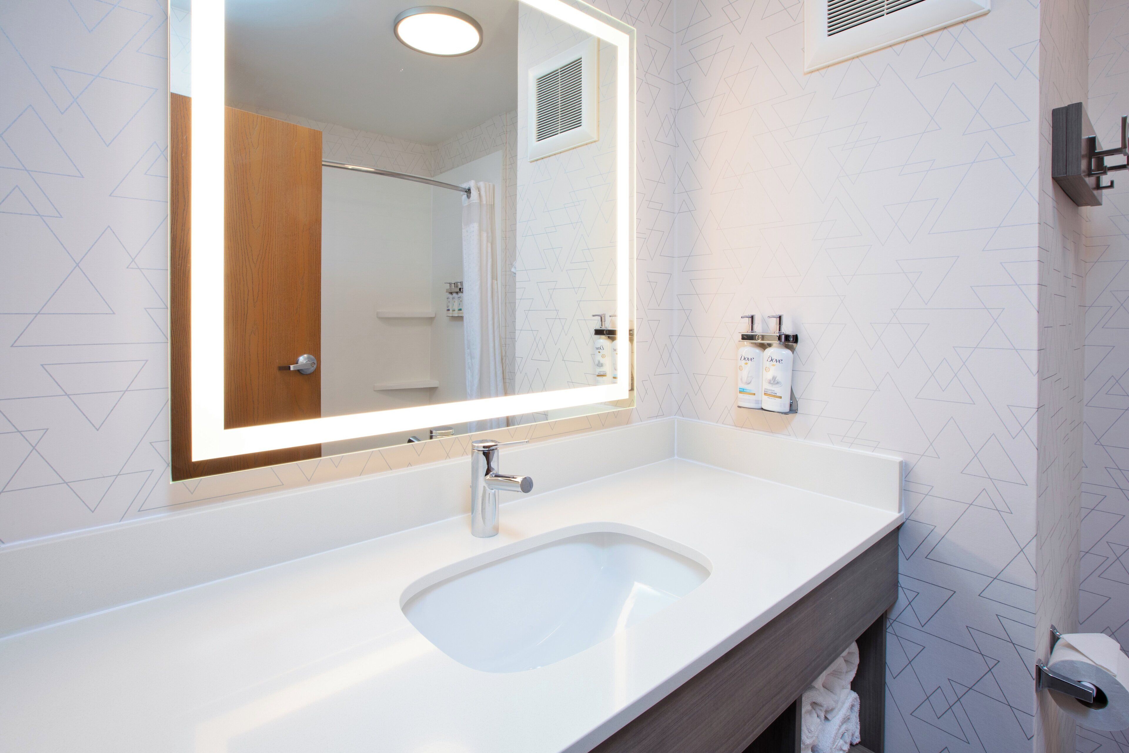 Un baño moderno con lavamanos blanco, un espejo grande y una puerta de madera.