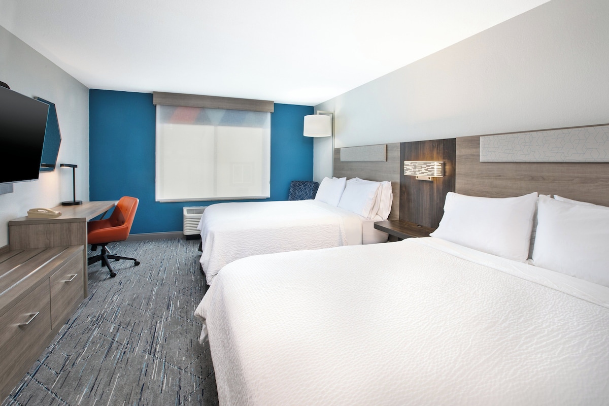 A hotel room with two beds, a desk, a chair, a TV, and a window.