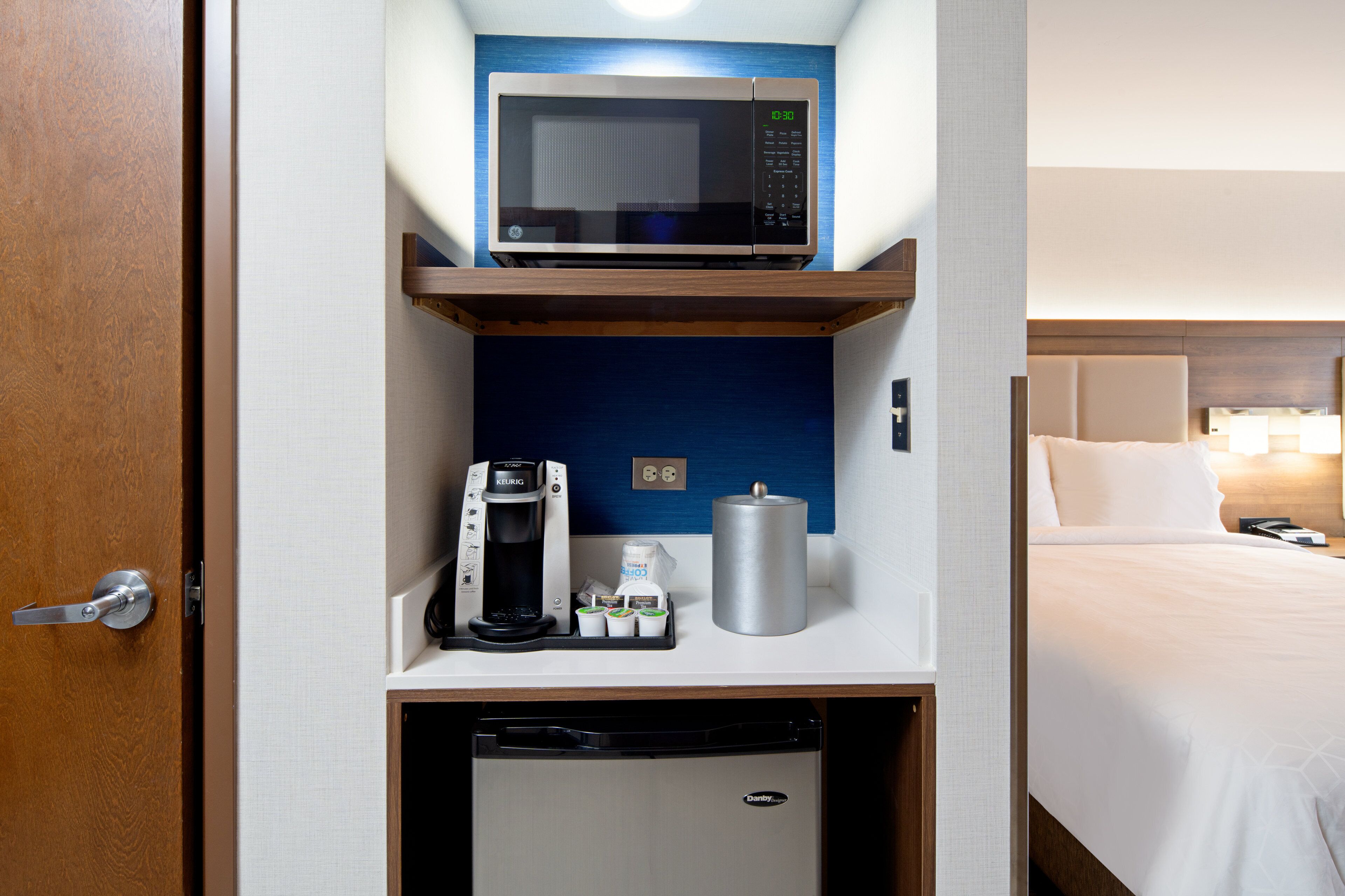 A compact hotel room with a microwave, coffee maker, and mini-fridge on a shelf, a bed with white linens, and a bedside table with a lamp.