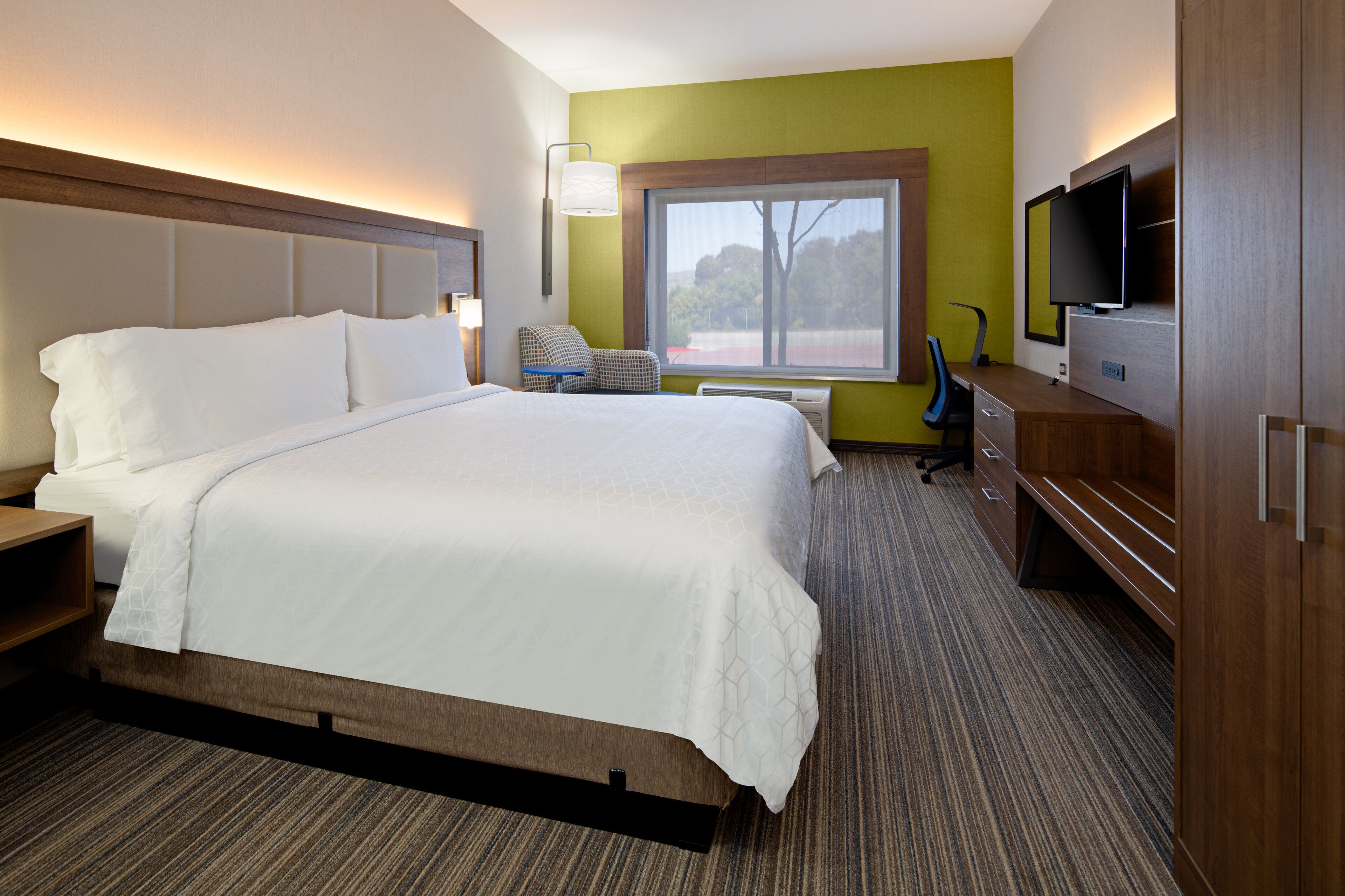 Une chambre d’hôtel avec un grand lit, un bureau avec une chaise, une télévision et une fenêtre donnant sur des arbres.