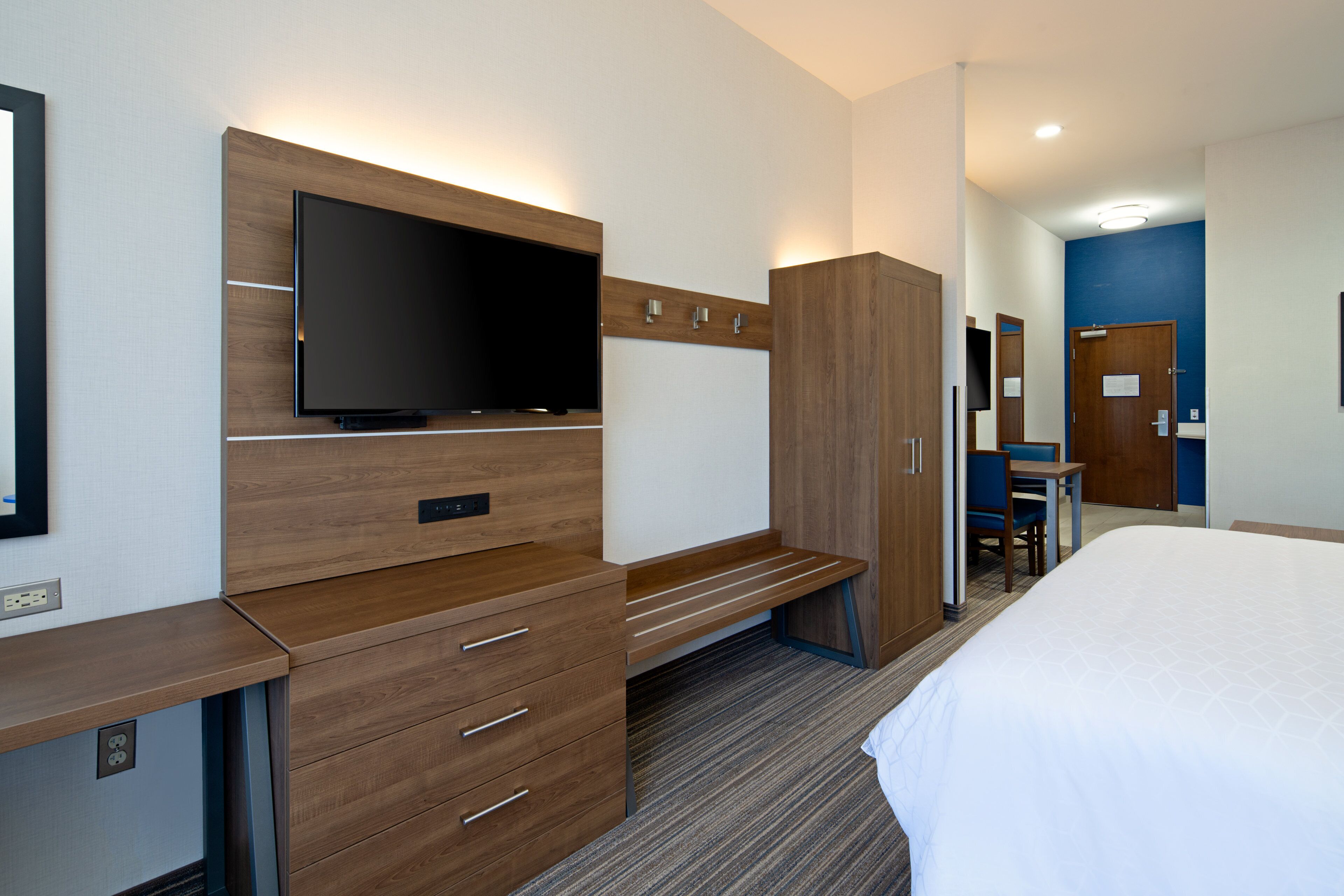 A hotel room with a bed, a desk with drawers, a flat-screen TV mounted on the wall, a wardrobe, and a mirror.