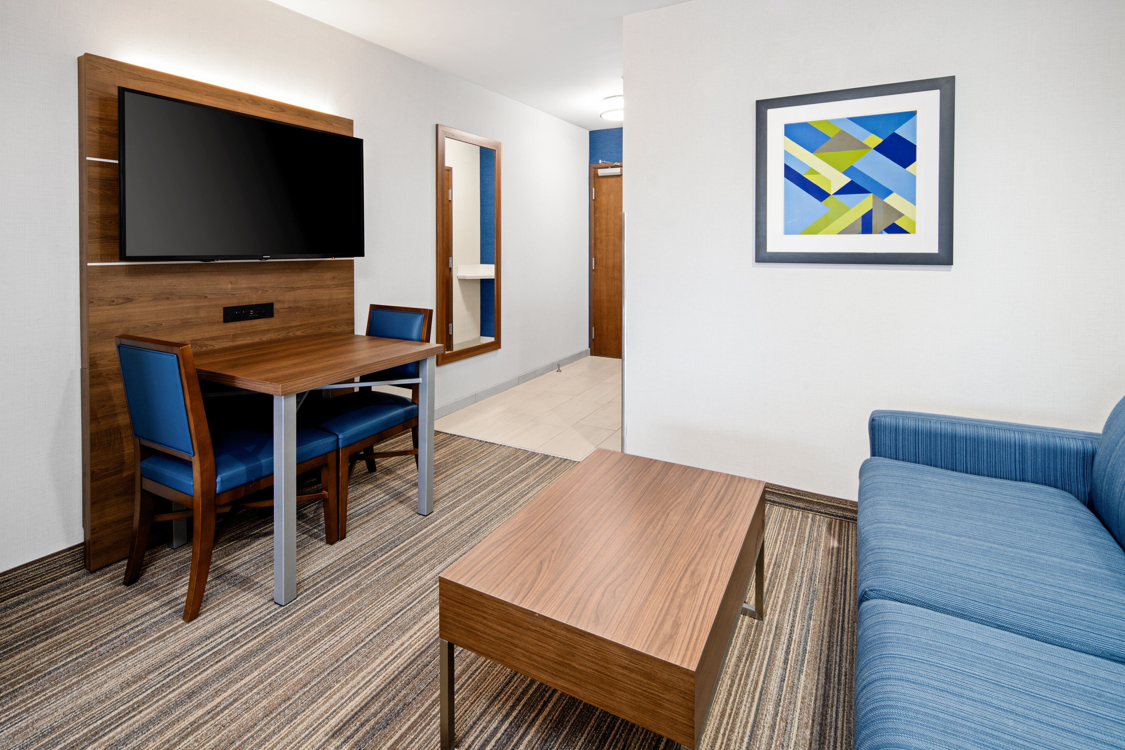 Una habitación de hotel moderna con una televisión de pantalla plana montada en un panel de madera, una mesa de comedor con sillas, un sofá azul y una obra de arte abstracta enmarcada en la pared.
