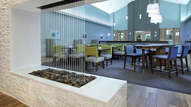 A modern hotel lobby with a stone wall, a seating area with tables and chairs, and a hanging light fixture.