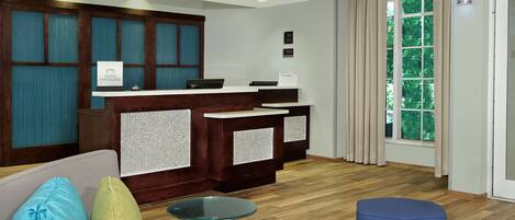 A hotel reception area with a wooden reception desk, a sofa with colorful cushions, a glass table, and a view of greenery through the window.