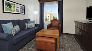 A hotel room with a dark blue sofa, a wooden coffee table, a brown leather ottoman, a brown armchair, a television, and a framed artwork on the wall.