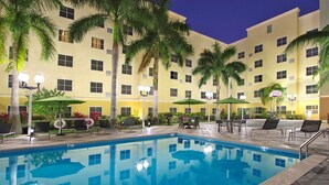 A swimming pool with lounge chairs and umbrellas, surrounded by palm trees and a multi-story building with balconies.