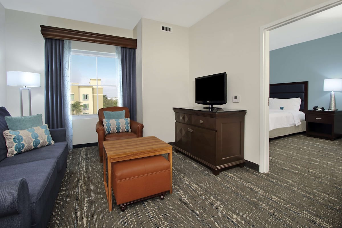 A hotel room with a sofa, a wooden table with an ottoman, a television on a cabinet, a bed with a nightstand, and a window with curtains.