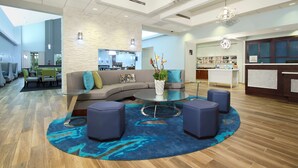 A modern hotel lobby with a curved sofa, round coffee table, and ottomans. There is a reception desk with a counter and shelves of amenities.