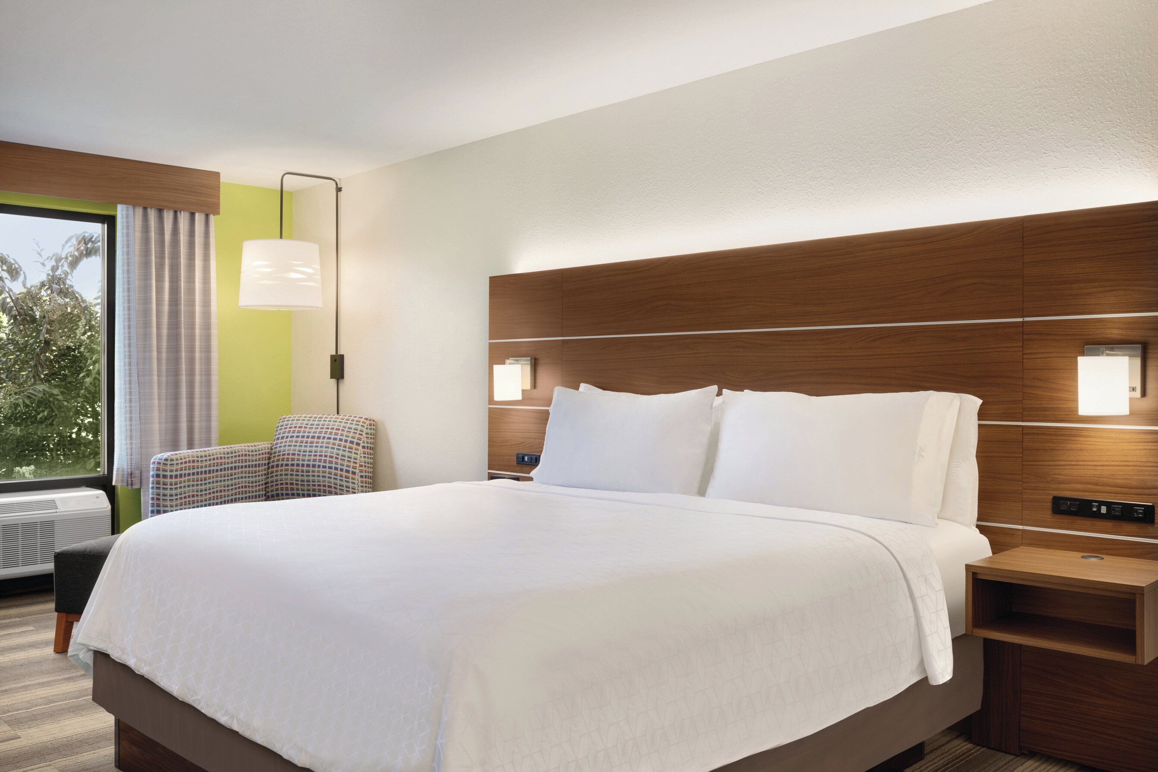 A hotel room with a large bed, a bedside table, a chair, and a window with curtains.