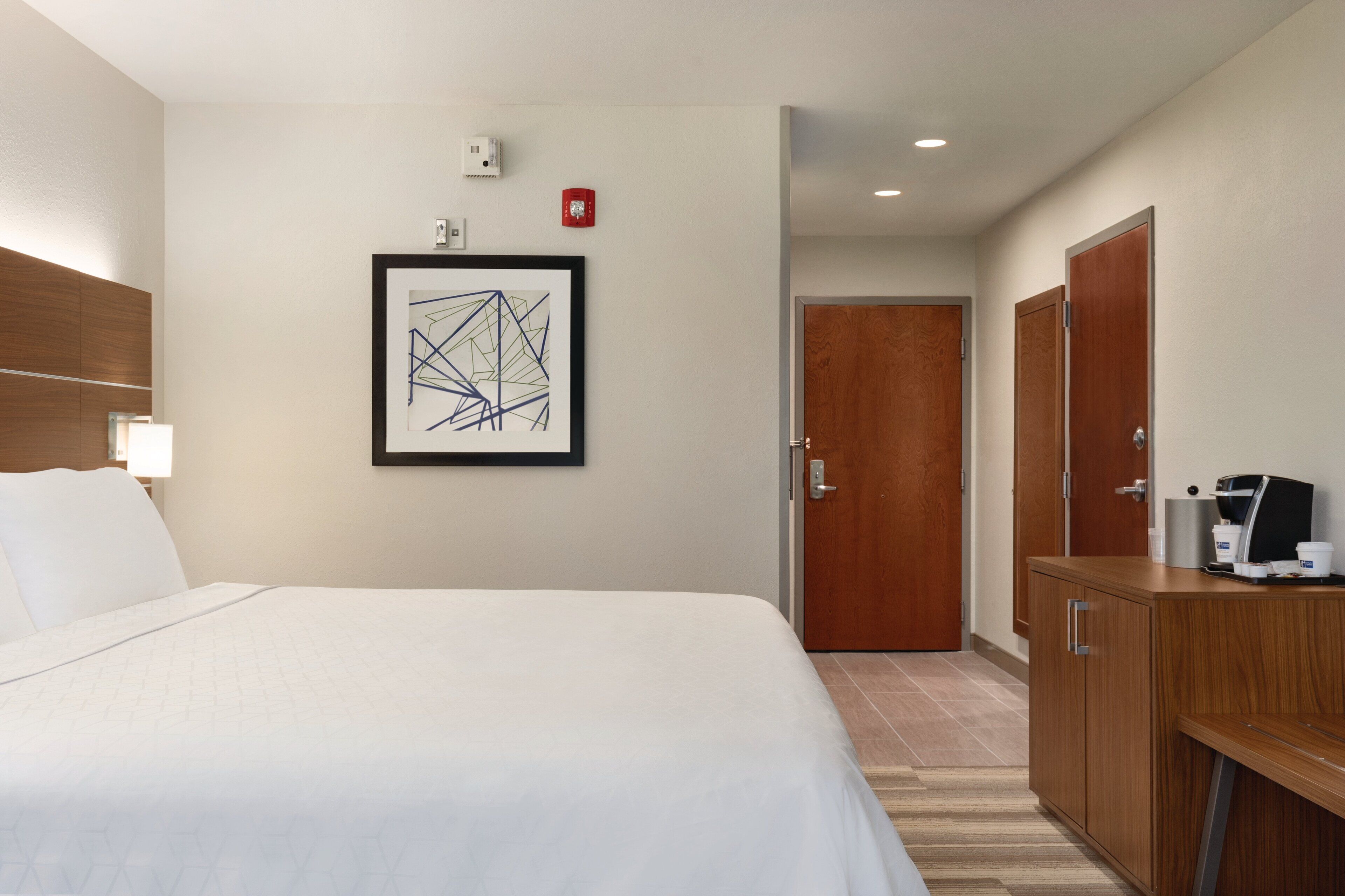 Habitación de hotel con una cama grande, un armario de madera, un cuadro abstracto enmarco y una máquina de café en la encimera.