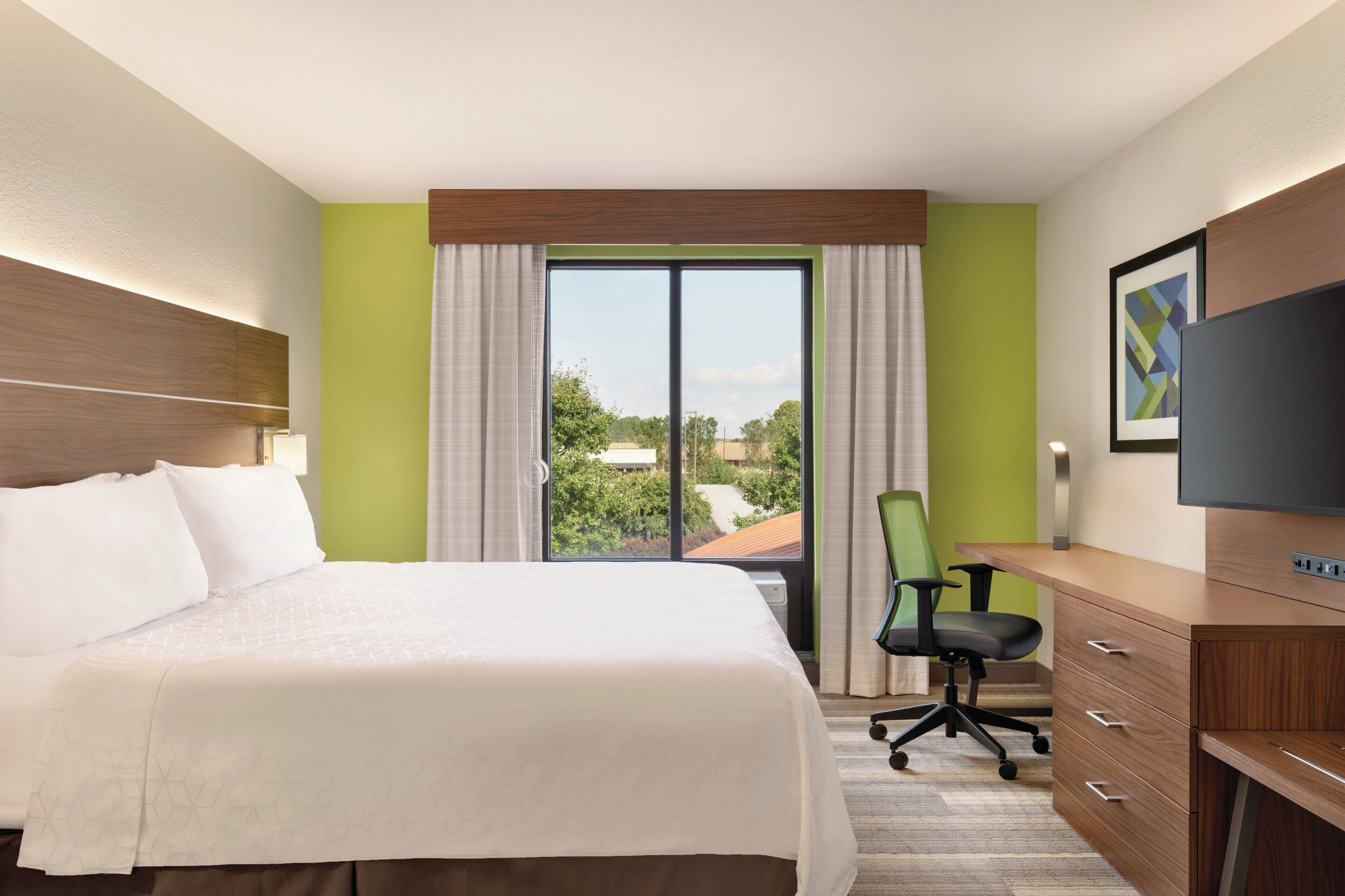 A hotel room with a bed, a desk, a chair, a television, and a window with a view of greenery.