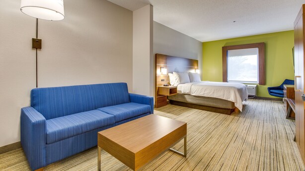 A hotel room with a blue sofa, a wooden coffee table, a bed with white bedding, a nightstand, a lamp, and a window with a view. - Holiday Inn Express Vincennes by IHG (Vincennes)