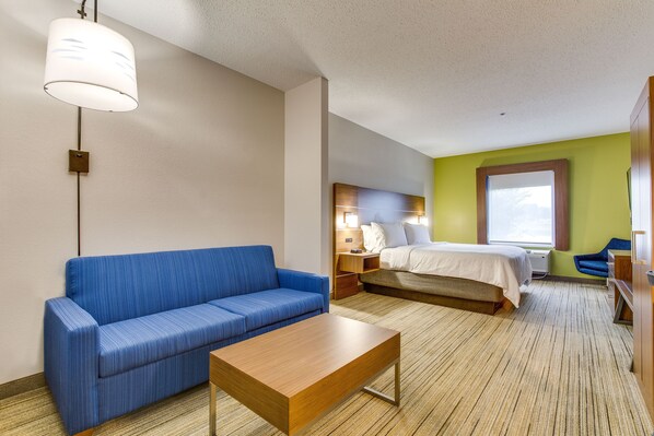 A hotel room with a blue sofa, a wooden coffee table, a bed with white bedding, a nightstand, a lamp, and a window with a view.